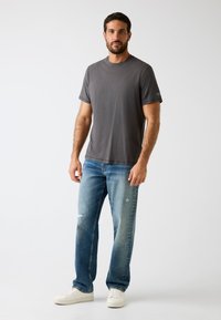 Graues Baumwoll-T-Shirt mit kurzen Ärmeln, kombiniert mit verwaschenen blauen Jeans mit geradem Bein und ausgefransten Details. Weiße Sneakers runden den Look ab.