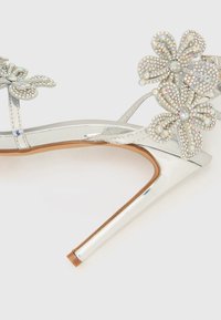 Ασημί πέδιλο με ψηλό τακούνι και γυαλιστερό φινίρισμα, με δύο διακοσμητικά λουλουδάκια στολισμένα με στρας. Ανοιχτό στ�ο δάκτυλο.