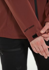 Röd vattentät jacka med en strukturerad tyg. Har en kardborreförslutning vid ärmen, svart sticksöm och etiketten "WPRO 10000" på ärmen.