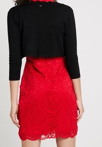 Schwarzes, kurzes Oberteil mit langen Ärmeln, kombiniert mit einem figurbetonten roten Spitzenkleid, das Blumemuster und einen welligen Saum aufweist.