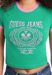 Žalias trumpas medvilninis marškinėliai su rožiniu apvadu ir centruojama grafika, kurioje užrašyta "GUESS JEANS" ir teniso raketės apvalios formos dizainu.