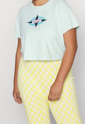 Persoon draagt een lichtblauw cropped t-shirt met grafische tekst en een gele hoge taille broek met wit hartjespatroon.