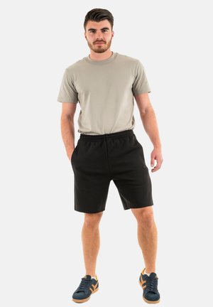 T-shirt khaki e pantaloni atletici neri. I pantaloni hanno una vita elasticizzata, vestibilità comoda e tasche laterali. Calzature: sneakers scure con accenti arancioni.