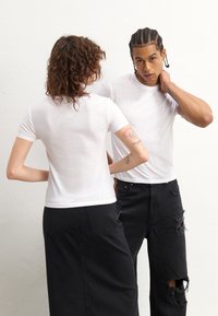 T-shirts brancos ajustados usados por duas pessoas, combinados com jeans pretos desgastados. O tecido parece ligeiramente transparente. Design simples, com mangas curtas.