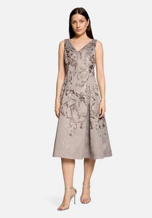 Kvinde iført ærmeløs lysegrå blomstret jacquard midikjole med V-udskæring og sølvfarvede højhælede sandaler, stående på hvid baggrund.