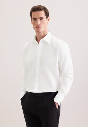 Mężczyzna ubrany w białą koszulę na długi rękaw z zapiętymi mankietami oraz czarne spodnie wizytowe, ręce w kieszeniach, neutralne tło.