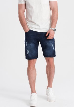 Pantalones cortos de denim en azul oscuro con detalles desgastados. Combinados con una camiseta clara y zapatillas blancas. Ajuste casual, justo por encima de la rodilla.