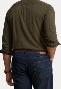 Oliwkowo-zielona koszula z długim rękawem z podwiniętymi mankietami, z tyłu z zakładką, w połączeniu z ciemnoniebieskimi dżinsami z kontrastowym przeszyciem i kieszeniami.