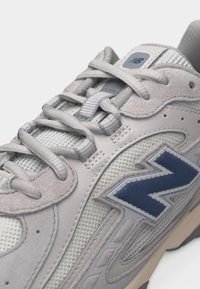 Κοντινό πλάνο μπεζ και γκρι αθλητικού παπουτσιού με μπλε λογότυπο New Balance, ύφασμα από πλέγμα, πάνελ από σουέτ και επίπεδοι κορδόνια δεμένα πάνω στη γλώσσα.