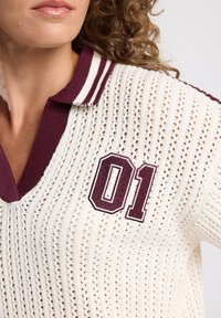 White knit polo with a v-neck, featuring maroon striped collar and embroidered number "01" in maroon. Textured, lightweight material.