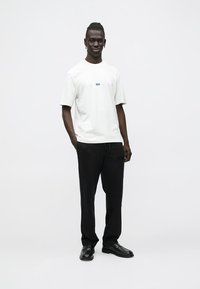 Bijela majica kratkih rukava s plavim logotipom, uparen s crnim hlačama nalik pidžami s natpisom "HUGO". Model stoji na jednobojnoj pozadini.