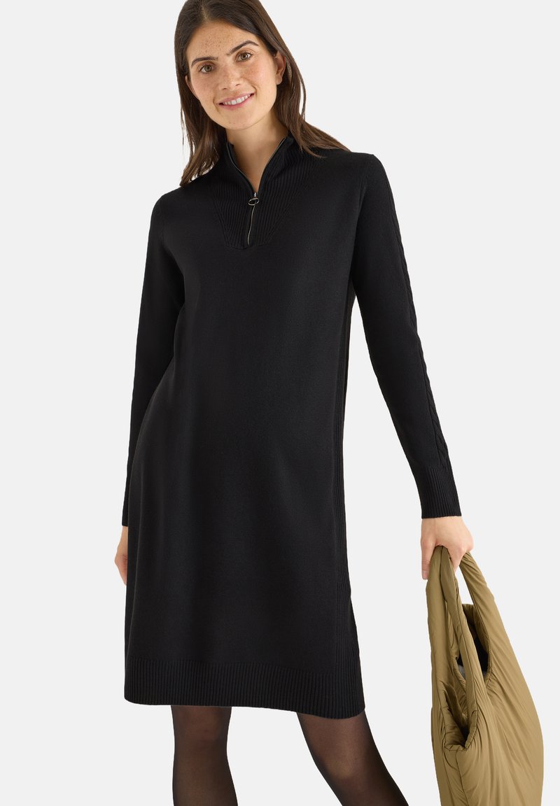Abito nero in maglia con colletto a metà zip, texture a costine, maniche lunghe e taglio dritto. La modella tiene una borsa leggera di colore verde oliva.