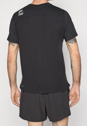 Man draagt een zwart T-shirt met de tekst "World Series of Fitness Racing" op de bovenrug en zwarte korte broek, met tatoeages op armen en benen.