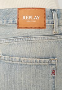 Šviesiai mėlyni džinsai su rudos odos etiketėmis, ant kurių užrašyta "REPLAY" ir "SINCE 1981". Yra raudonas siuvinėjimas ant užpakalinio kišenės.