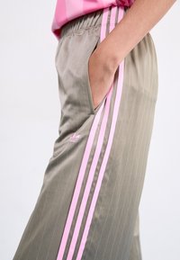 Grå träningsbyxor med ränder, med rosa sidosträck och en Adidas-logotyp. Elastisk midja och sidofickor är synliga.