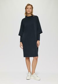Marineblauwe oversized jurk met hoge kraag en vleermuismouwen, gemaakt van zachte stof. Gedragen met witte platform sneakers.