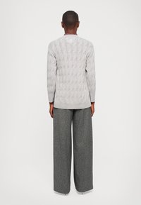Woman standing facing away, wearing light gray cable-knit sweater and wide-leg dark gray pants against plain white background.