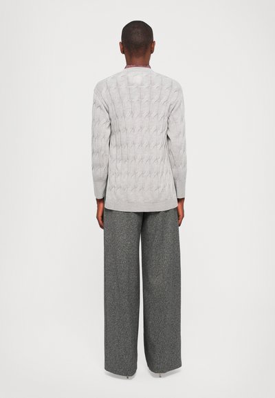 Donna in piedi di spalle, che indossa un maglione grigio chiaro a trecce e pantaloni larghi grigio scuro, su sfondo bianco semplice.