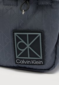 Τσάντα Calvin Klein σε σκούρο γκρι ύφασμα με σχεδιασμό με μοτίβο, λαστιχένιο λογότυπο και υφασμάτινες λεπτομέρειες. Κλείσιμο με ρυθμιζόμενο κορδόνι.