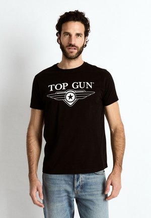 Mand med krøllet hår og skæg iført en sort Top Gun T-shirt og blå jeans, stående mod en ensfarvet hvid baggrund.