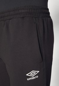 Čierne tepláky vyrobené z mäkkého materiálu s elastickým pásom, so značkou Umbro v bielej farbe na spodnej strane.