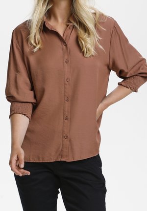 Kobieta ubrana w luźną brązową koszulę na guziki z marszczonymi mankietami i czarne spodnie, stojąca z jedną ręką na biodrze, a drugą swobodnie opuszczoną.