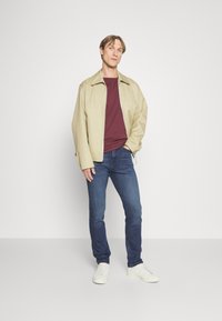 Beige leichte Jacke, kastanienbraunes Hemd, blaue Jeans und weiße Sneakers. Die Jacke hat einen Reißverschluss, eine lockere Passform und eine weiche Textur.