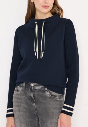 Marineblå pullover med høj krave og snore, med ribstrikkede ærmer med stribede detaljer, kombineret med grå denimjeans.
