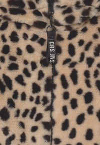 Leopardmönstrad fuskpäls med svarta fläckar på en beige bakgrund. Har en svart dragkedja med en vit text. Mjuk textur.