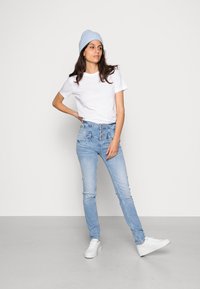 Denim de cintura alta azul claro com cinco botões à frente, bolsos padrão e um corte justo. Combinado com uma t-shirt branca e um gorro azul claro.