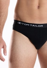 Black cotton brief featuring a logoed elastic waistband, smooth texture, and a fitted design, positioned on a model's lower body.