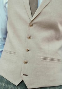 Beige Leinenweste mit fünf Holzknöpfen und einer kleinen Tasche. Sichtbare Akzentnähte. Kombination mit einer strukturierten braunen Krawatte.