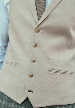 Gilet en lin beige avec cinq boutons en bois et une petite poche. Couture d'accent visible. Association avec une cravate marron texturée.
