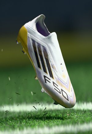 White soccer boot with yellow and purple accents, featuring a textured upper, three black stripes, and molded studs on a grassy surface.