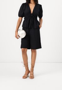 Top nero con maniche a sbuffo e scollatura a V profonda, abbinato a pantaloni corti neri e sandali beige con lacci. Tiene una borsa bianca.