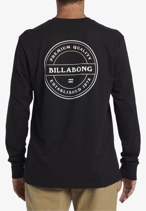 Mężczyzna w czarnej koszulce z długim rękawem z okrągłym logo z napisem "Premium Quality Billabong Established 1973" na plecach.