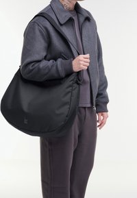 Grande borsa a tracolla in nylon nero con una texture liscia e una singola tracolla, tenuta da una persona che indossa una giacca grigia con cerniera e pantaloni.