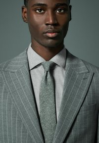 Chaqueta de traje a rayas grises con corbata a juego; camisa gris clara por debajo. Textura suave; corte ajustado; solapas estrechas y detalle de bolsillo en el pecho.