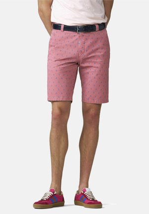 Man die roze korte broek met patroon, marineblauwe riem, wit T-shirt en rode sneakers met blauwe strepen draagt, staande tegen een effen achtergrond.