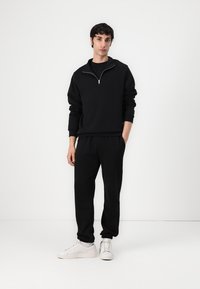 Sudadera con cremallera negra y joggers a juego, hechos de una tela texturizada. Combinados con zapatillas blancas, de pie frente a un fondo blanco liso.