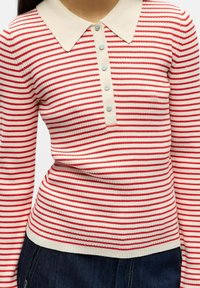 Raudonai ir baltai dryžuotas megztinis su smėlio spalvos apykakle, turintis sagų skyrių ir riboto rašto tekstūrą. Dėvimas su tamsiomis džinsinėmis kelnėmis.