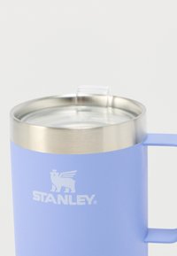 Tasse en acier inoxydable avec finition mate bleu clair, couvercle transparent et poignée latérale. Présente un logo blanc en relief sur le devant.