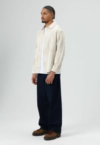 Crèmekleurig, gestructureerd overhemd met knopen en een brede kraag, gecombineerd met een wit T-shirt en donkerblauwe, wijde broekspijpen. Bruine wandelschoenen.