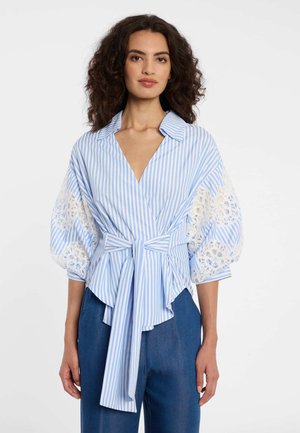 Vrouw draagt een blauw-wit gestreepte blouse met kanten balloonmouwen en een ceintuur in de taille, gecombineerd met donkerblauwe broek, staand tegen een witte achtergrond.