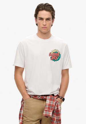 T-shirt bianca in cotone con logo "Super Dry" multicolore sul petto sinistro. Il modello indossa pantaloni khaki e una camicia a quadri rossa legata in vita.