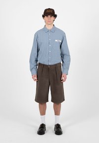 Jeune homme debout portant une chemise à rayures bleues avec boutons, un short marron jusqu'aux genoux, des chaussettes blanches, des chaussures noires et une casquette foncée avec un texte.