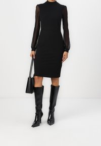 Schwarzes Kleid mit Rundhalsausschnitt und transparenten langen Ärmeln, kombiniert mit knielangen schwarzen Stiletto-Stiefeln und einer kleinen schwarzen Handtasche.