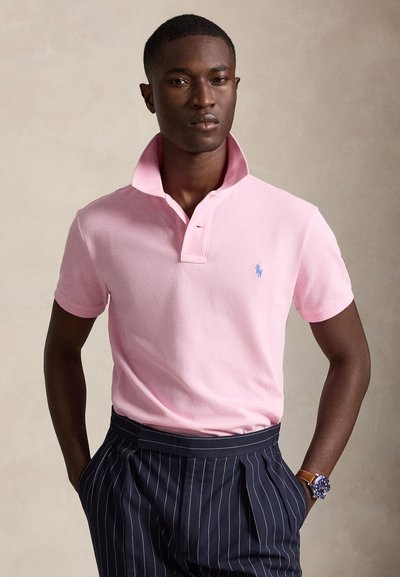 Giovane uomo che indossa una polo rosa chiaro con il colletto alzato, pantaloni blu navy a righe sottili e un orologio con cinturino in pelle marrone.