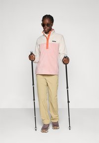 Persona che indossa occhiali da sole, un pullover di pile rosa e bianco, pantaloni beige e scarpe da trekking, mentre tiene in mano due bastoni da trekking neri.