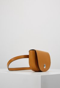 Torba na ramię z jasnobrązowej skóry z klapką zapinającą i okrągłym metalowym akcentem. Posiada cienki regulowany pasek i gładką fakturę powierzchni.